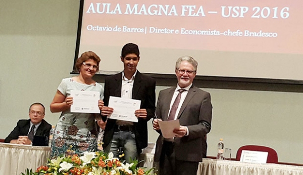 Ao lado da professora Izabel Gil, aluno Guilherme Augusto Santana (centro) exibe prêmio  recebido durante aula magna dos cursos de graduação da FEA-USP (Imagem: Divulgação).